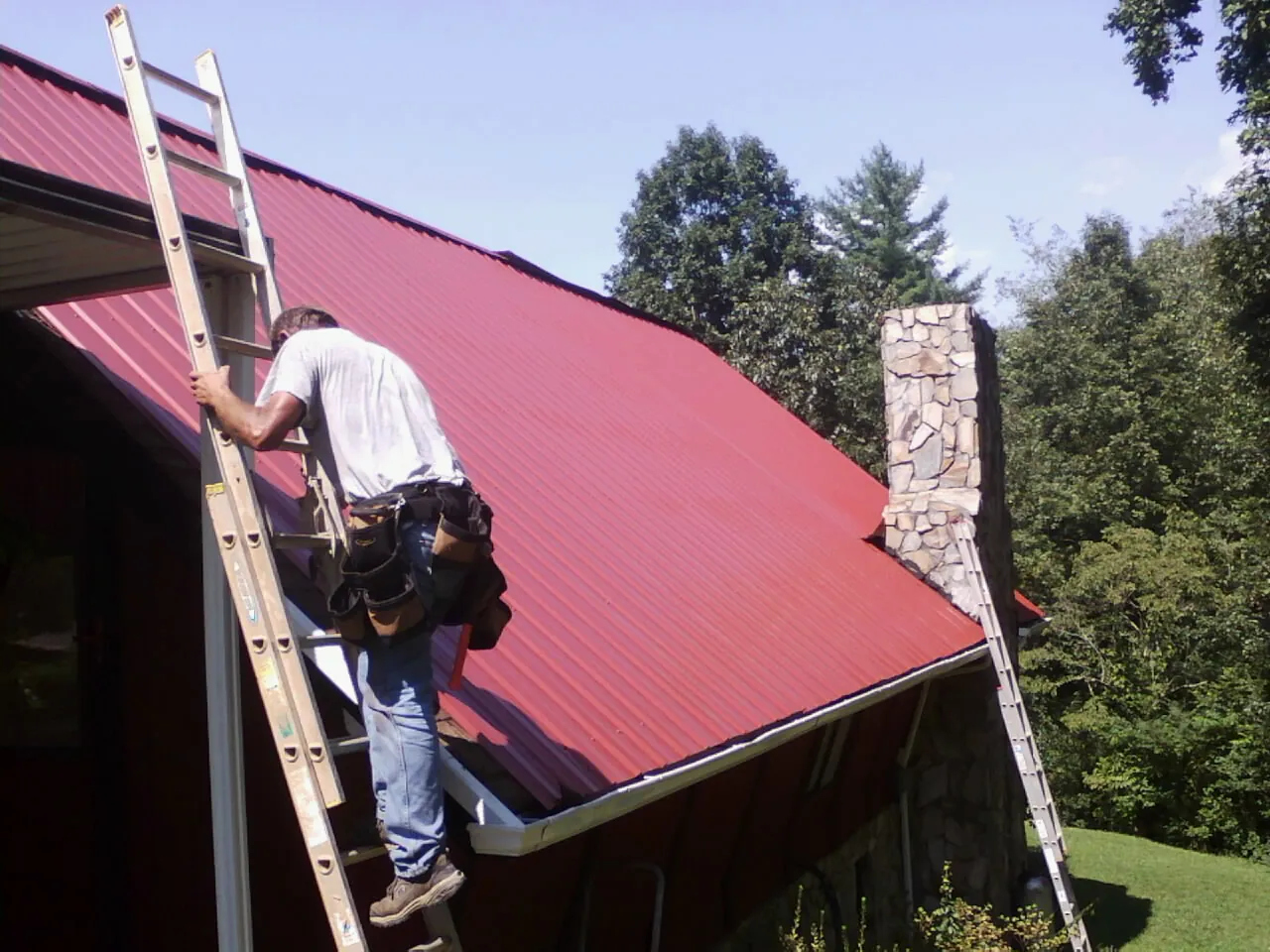 Roofer on a ladder beside a red metal roof and stone chimney — Soffit & Fascia Repair by Roofing Brothers in Absaraka